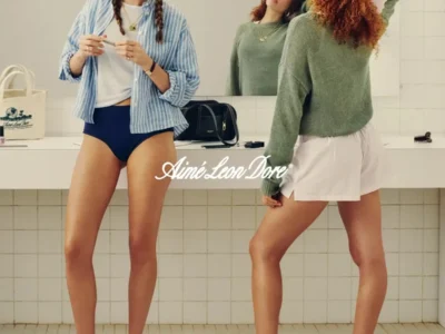 Two models in a dressing-room setting wearing Aimé Leon Dore spring essentials, including striped shirts, knitwear, briefs, shorts, and ballet flats