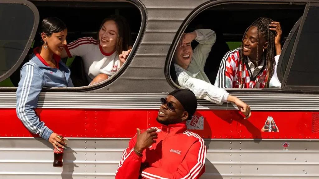 Friends wearing Adidas Originals x Coca-Cola FIFA World Cup 2026 apparel gather around a vintage bus, showcasing red-and-white sportswear, football-inspired jerseys, and collaborative branding in a nostalgic campaign scene