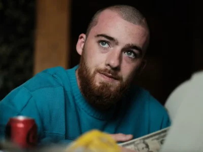Close-up portrait of Angus Cloud as Fezco in Euphoria, wearing a blue knit sweater while seated indoors, with cash and a soda can visible in the foreground, capturing the character’s thoughtful and understated presence