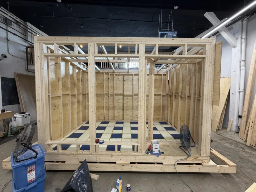 Wooden framing of the floating convenience store under construction inside a workshop, showing the structural skeleton, open wall studs, and checkerboard flooring before exterior finishes were install