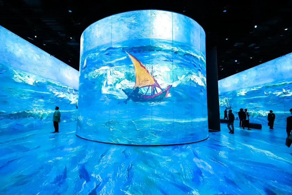 Visitors stand inside an immersive digital gallery where towering curved screens display a vivid blue seascape with a sailboat, surrounding the room in animated brushstroke