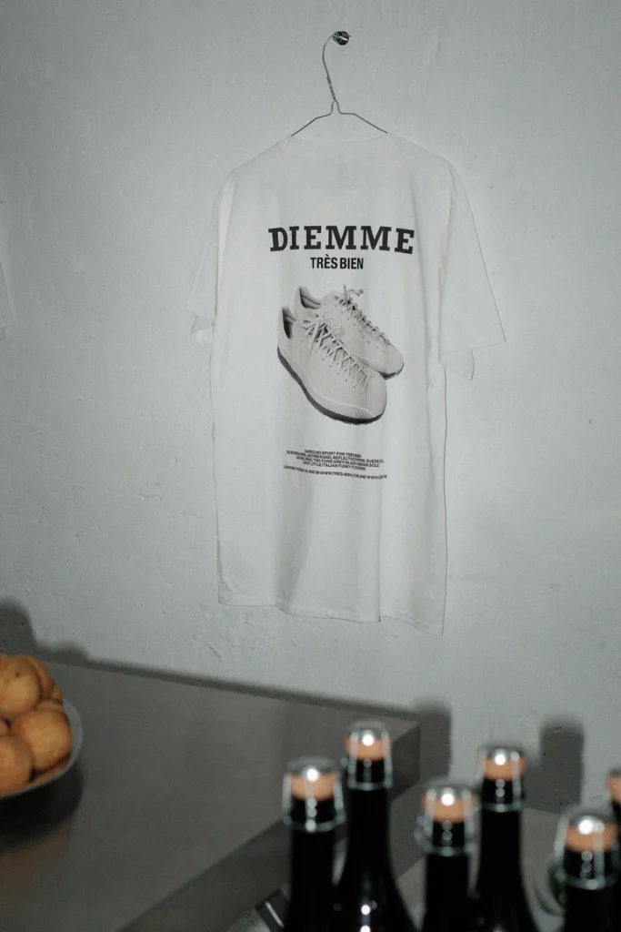 White DIEMME × Très Bien graphic T-shirt hanging on a metal hook against a textured wall, featuring a large sneaker print on the back above a table with candles and a bowl of fruit