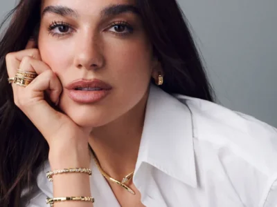 Close-up portrait of a Dua Lipa with long dark hair wearing a crisp white button-up shirt and layered gold jewelry. They rest their face on one hand while looking directly at the camera with a neutral expression. Multiple gold rings, hoop earrings, bracelets, and a delicate pendant necklace complete the polished look against a soft gray studio background