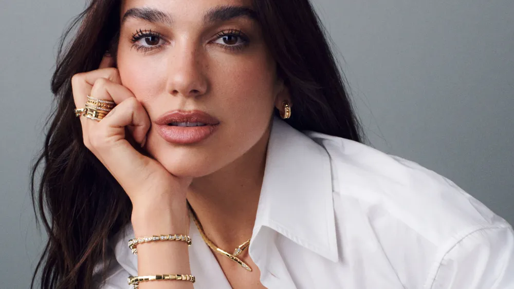 Close-up portrait of a Dua Lipa with long dark hair wearing a crisp white button-up shirt and layered gold jewelry. They rest their face on one hand while looking directly at the camera with a neutral expression. Multiple gold rings, hoop earrings, bracelets, and a delicate pendant necklace complete the polished look against a soft gray studio background