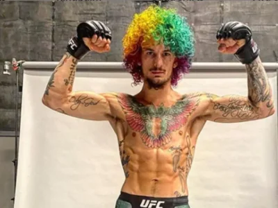 Shirtless, heavily tattooed UFC fighter flexing powerfully with both arms raised, wearing a vibrant rainbow curly wig, black MMA gloves, and branded UFC shorts. Studio backdrop with concrete walls and lighting rig. Bold, playful, and energetic pose blending combat sports with colorful street style