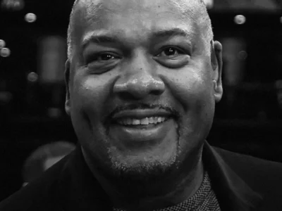 Close-up black-and-white portrait of a smiling man wearing a dark coat, photographed indoors against a softly blurred background with lights and reflections