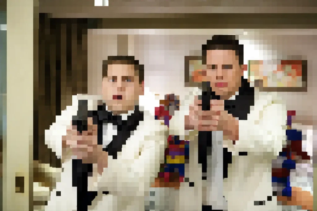 pixelated still image showing two men in white tuxedo jackets and black bow ties standing side by side in a hotel-style room. Both appear to be aiming handguns toward the camera, though facial details and objects are obscured by large mosaic-style pixels. Warm indoor lighting, framed wall art, and colorful decorative figures are visible in the blurred background