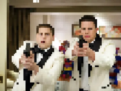 pixelated still image showing two men in white tuxedo jackets and black bow ties standing side by side in a hotel-style room. Both appear to be aiming handguns toward the camera, though facial details and objects are obscured by large mosaic-style pixels. Warm indoor lighting, framed wall art, and colorful decorative figures are visible in the blurred background
