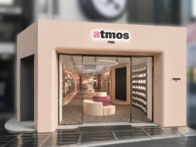 Exterior storefront of atmos Pink featuring a minimalist cream façade, illuminated atmos Pink logo above a full-height glass entrance, sculptural white and pink display fixtures visible inside, mannequin displays, walls of sneakers, and contemporary curved lighting that extends throughout the boutique interior