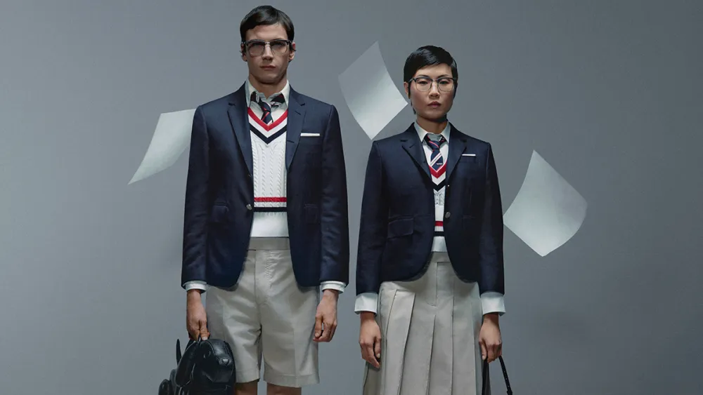 Studio campaign image featuring two models in preppy navy blazers, striped V-neck knitwear, pleated shorts and skirts, ties, and eyeglasses against a minimalist gray backdrop with floating sheets of paper