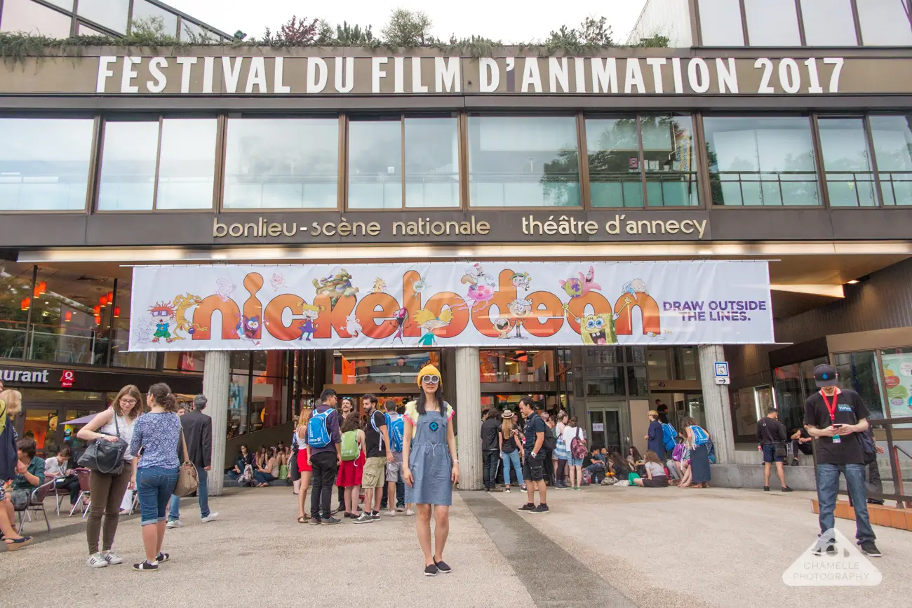 Crowds gather outside the Bonlieu Scène Nationale in Annecy, France, during the 2017 Annecy International Animation Film Festival. Large signage spans the building façade, while a colorful Nickelodeon banner featuring animated characters hangs above the entrance. Festival attendees stand, chat, and queue outside the venue, with one person posing in the center of the plaza