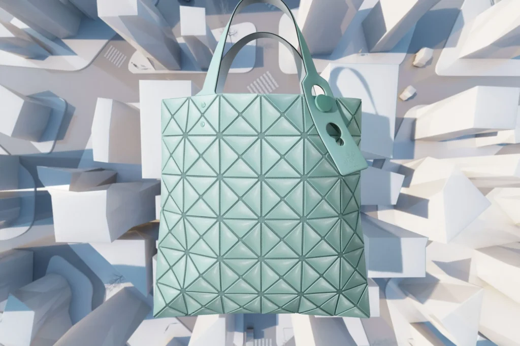 Mint-green geometric tote bag from BAO BAO ISSEY MIYAKE floats above a stylized miniature cityscape, showcasing the brand’s signature triangular panel construction. The futuristic composition highlights the bag’s architectural design and sculptural form