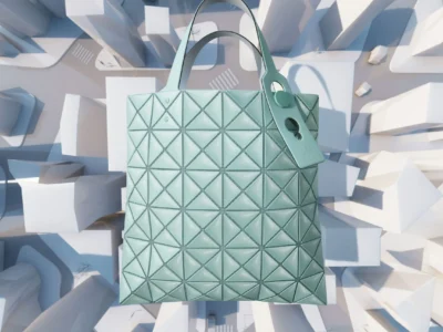 Mint-green geometric tote bag from BAO BAO ISSEY MIYAKE floats above a stylized miniature cityscape, showcasing the brand’s signature triangular panel construction. The futuristic composition highlights the bag’s architectural design and sculptural form