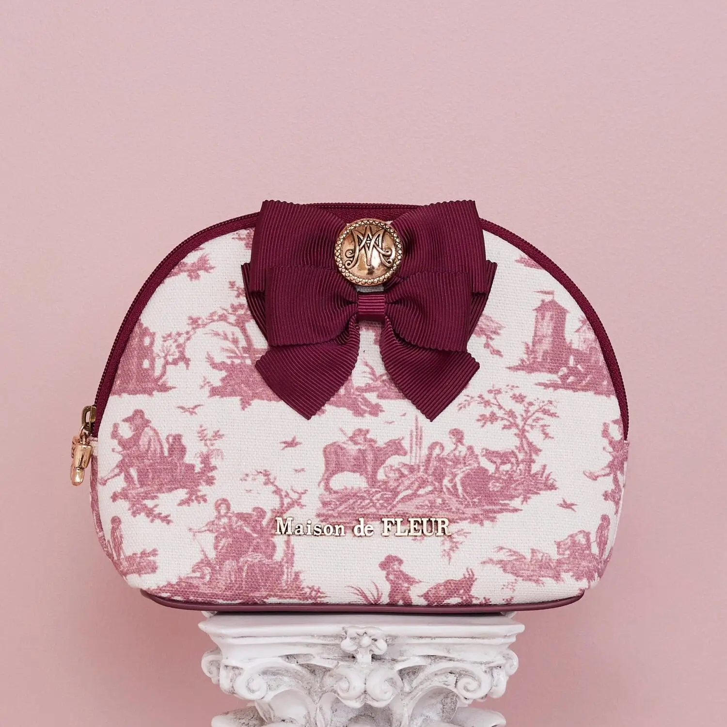 Small dome-shaped cosmetic pouch in cream canvas with a dusty rose toile pastoral print. The pouch features burgundy trim, a matching grosgrain ribbon bow with a gold-tone medallion at the center, and metallic logo lettering across the front. Displayed atop an ornate white decorative pedestal against a soft blush-pink background, the design evokes vintage French elegance