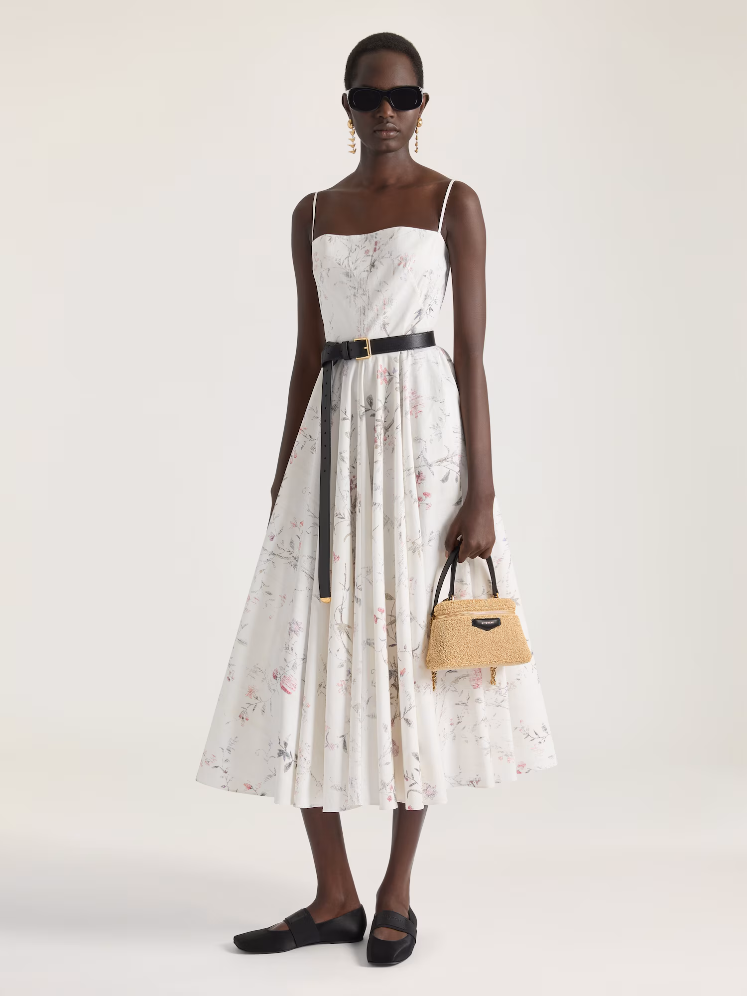 Model wearing a white floral-print midi dress with delicate spaghetti straps, styled with a black leather belt, flat black shoes, oversized sunglasses, and a compact raffia-textured Givenchy handbag