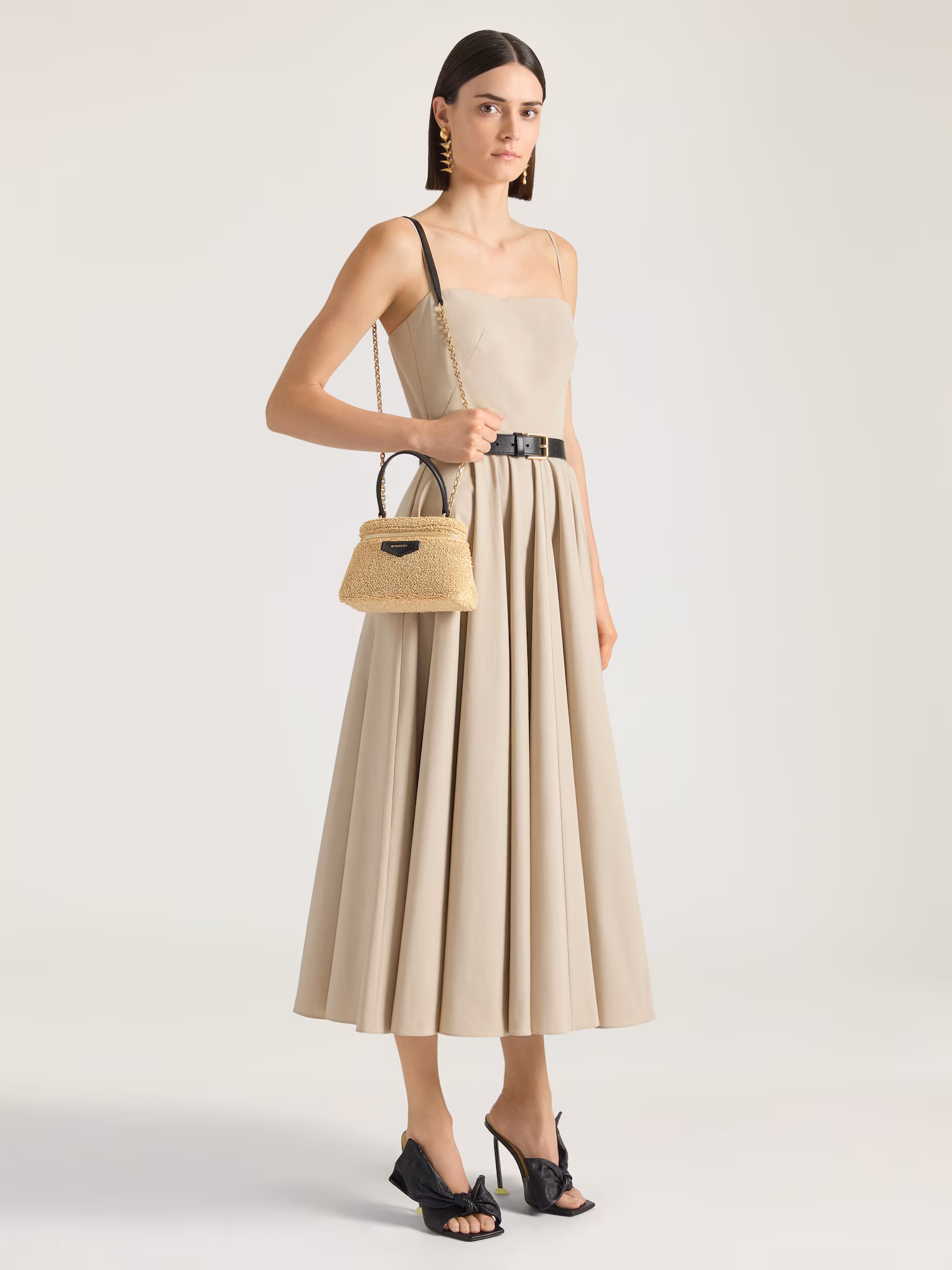 Model in a minimalist beige midi dress featuring a fitted bodice and full pleated skirt, accessorized with a black belt, sculptural earrings, high-heeled sandals, and a raffia Givenchy top-handle bag