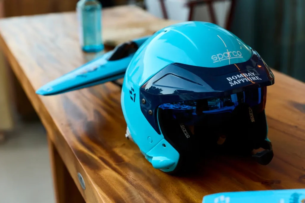 A custom Sparco racing helmet featuring Bombay Sapphire branding sits on a polished wooden table beside a streamlined model powerboat. The scene highlights precision engineering, motorsport aesthetics, and luxury partnership