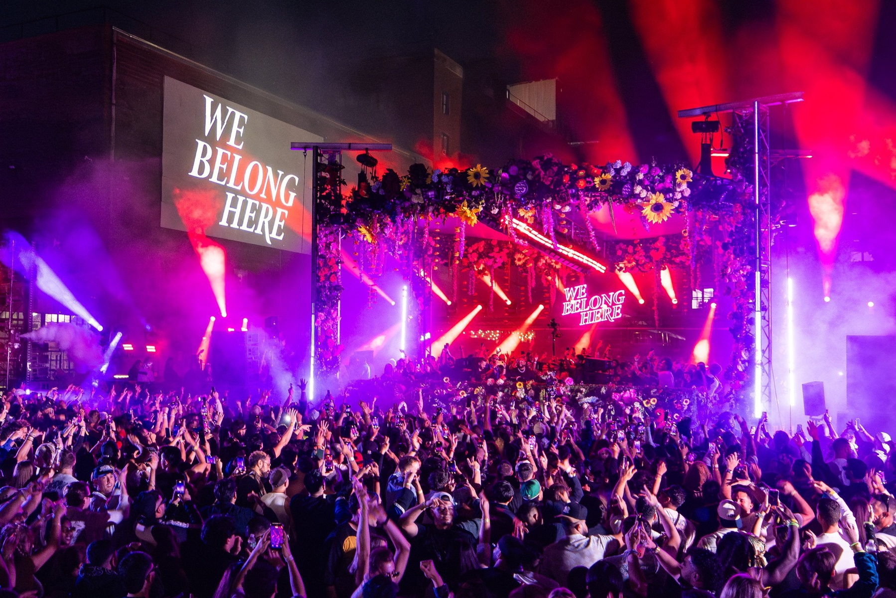 Large outdoor music festival at night with a packed crowd facing a flower-covered stage illuminated by vibrant pink, purple, and red lights. Massive screens display the message “WE BELONG HERE” as attendees raise their hands and phones in celebration