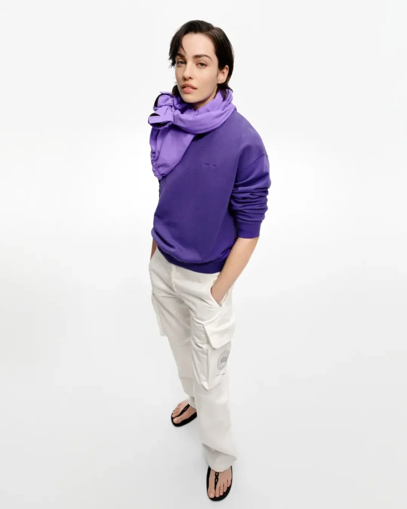 Model wearing a vibrant purple crewneck sweatshirt, matching scarf, and white utility cargo trousers from the Canada Goose Summer 2026 collection