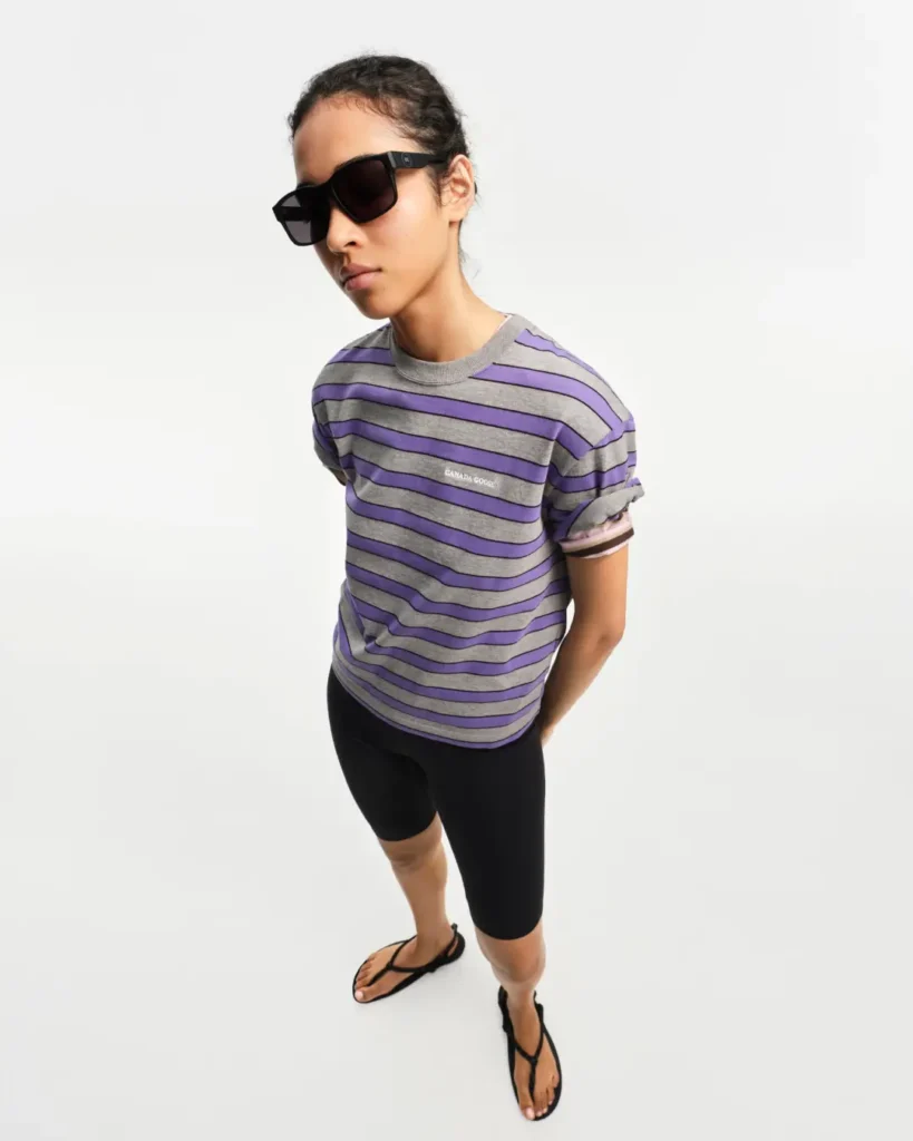 Model styled in a grey and purple striped Canada Goose T-shirt, cycling shorts, and oversized sunglasses for a casual summer look