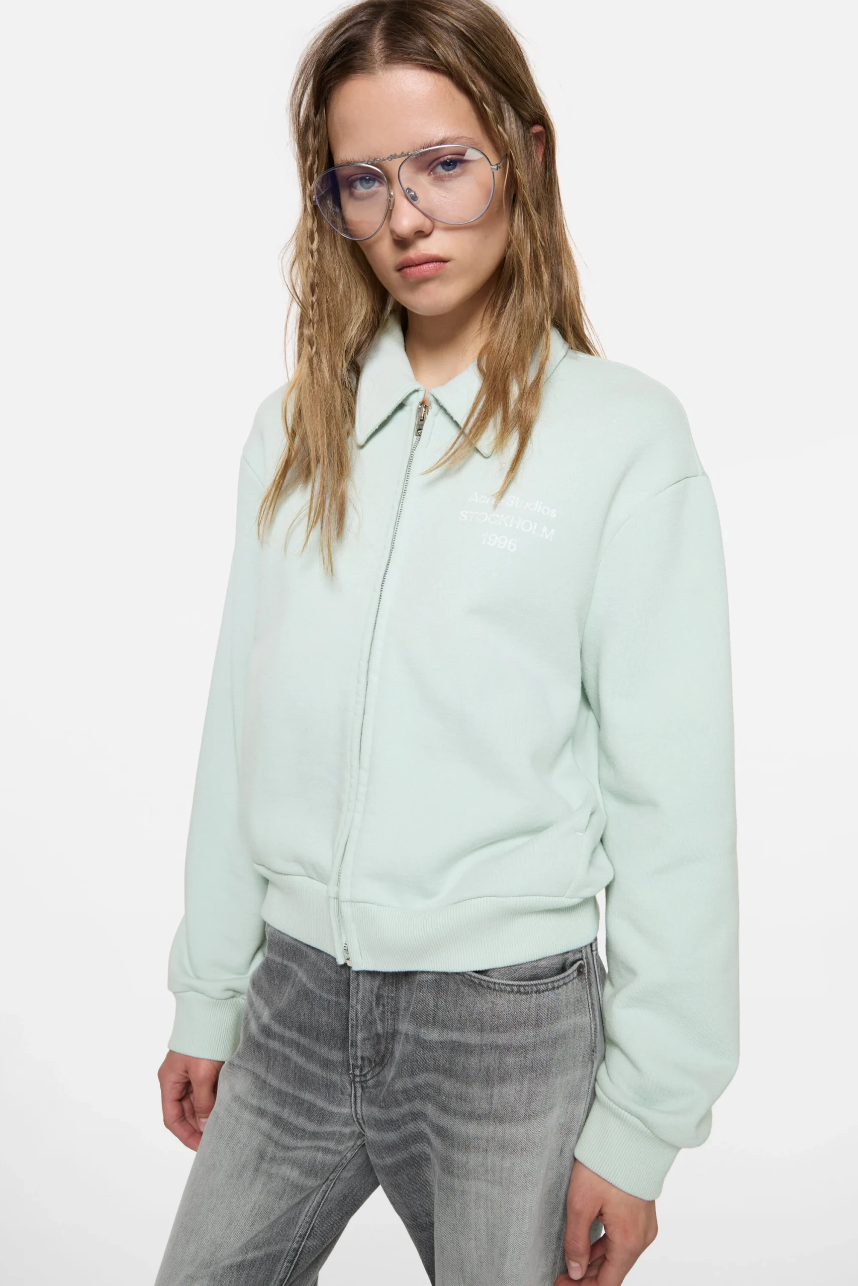 Model wearing a pale mint Acne Studios zip-up sweatshirt jacket with a pointed collar, subtle Stockholm 1996 logo print, oversized fit, grey washed denim jeans, and oversized aviator-style glasse