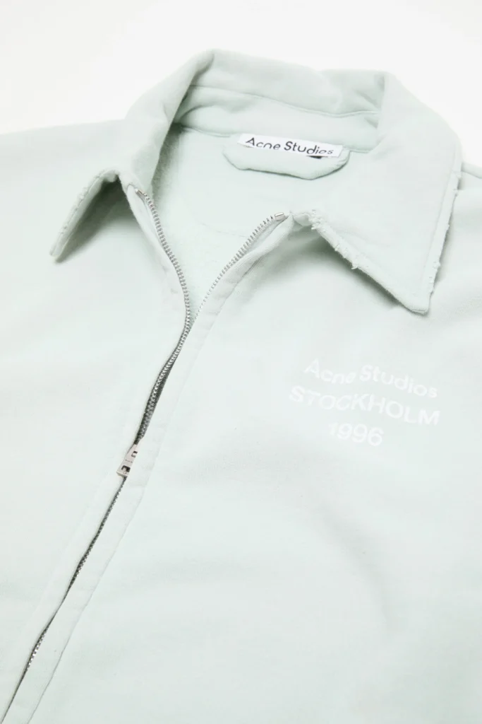 Close-up detail of the Acne Studios mint green zip-up sweatshirt jacket showcasing the pointed collar, silver zipper closure, soft fleece fabric, and tonal Stockholm 1996 chest brand