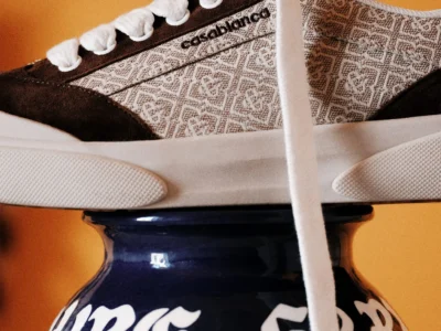 Close-up side profile of a Casablanca Del Mar shoe in a brown monogram colorway, featuring patterned textile panels, dark suede overlays, thick white laces, and a cream sole, balanced atop a blue ceramic vase against a warm amber backdrop