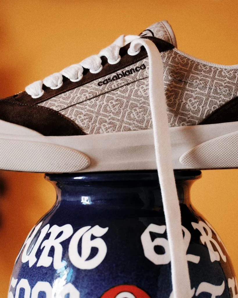 Close-up side profile of a Casablanca Del Mar shoe in a brown monogram colorway, featuring patterned textile panels, dark suede overlays, thick white laces, and a cream sole, balanced atop a blue ceramic vase against a warm amber backdrop