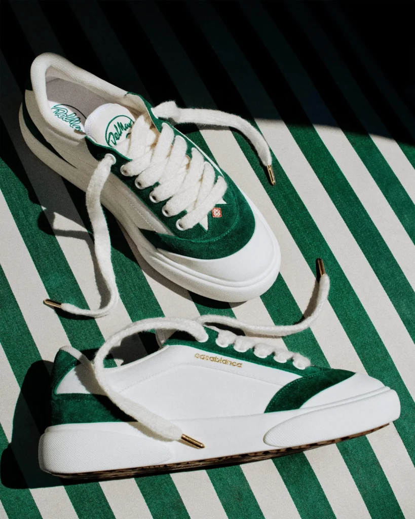 Two Casablanca Del Mar sneakers in a white-and-green colorway arranged on a green-and-white striped surface, showcasing oversized fuzzy laces, suede accents, and gold-tipped lace ends under dramatic sunlight