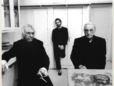 Black-and-white portrait of three men dressed in dark clothing inside a modest, vintage-style kitchen. Two older men sit in the foreground with serious expressions while a younger man stands between them in the background, leaning against white cabinet doors. The image features stark contrast, worn surfaces, and a quiet, cinematic atmosphere that emphasizes character, age, and presence