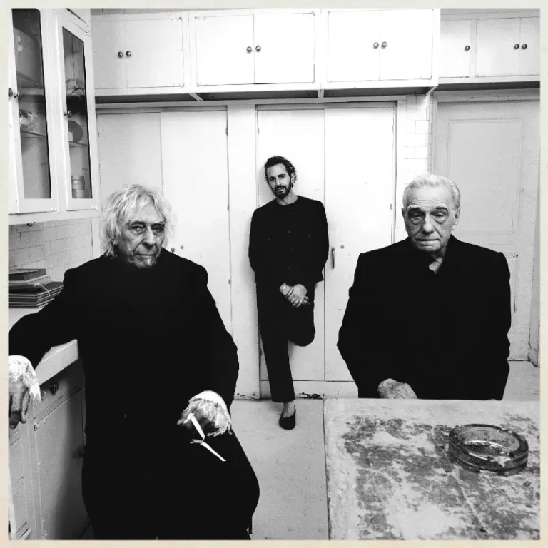 Black-and-white portrait of three men dressed in dark clothing inside a modest, vintage-style kitchen. Two older men sit in the foreground with serious expressions while a younger man stands between them in the background, leaning against white cabinet doors. The image features stark contrast, worn surfaces, and a quiet, cinematic atmosphere that emphasizes character, age, and presence