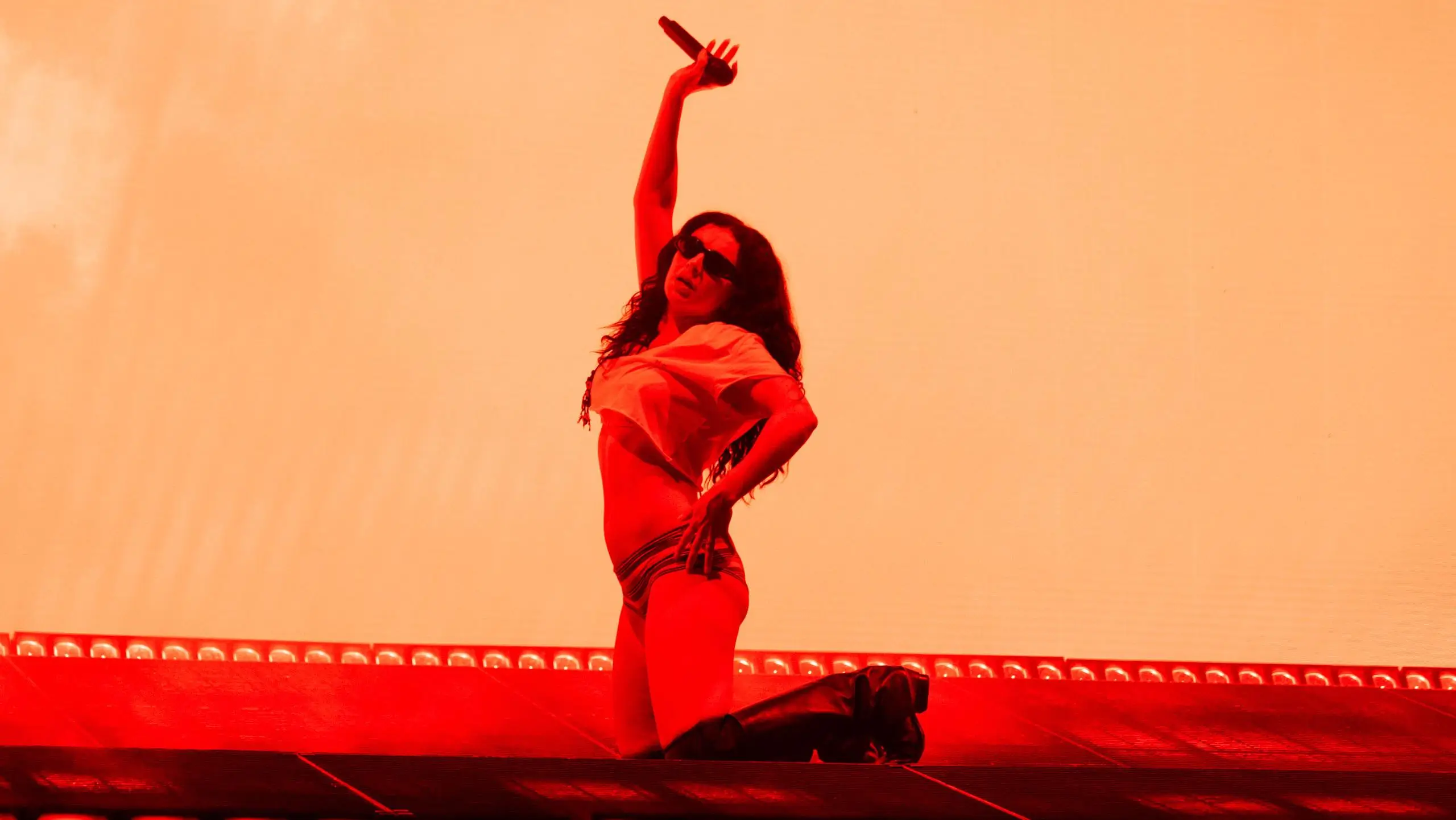 Charli XCX kneels onstage beneath intense red lighting, wearing sunglasses, a cropped white top, and high boots while striking a dramatic performance pose