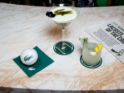 Marble-topped Club Lacoste bar display featuring two signature cocktails on branded coasters, a Lacoste-logo tennis ball on a green napkin, and a newspaper-style menu, blending tennis-inspired luxury with refined hospital