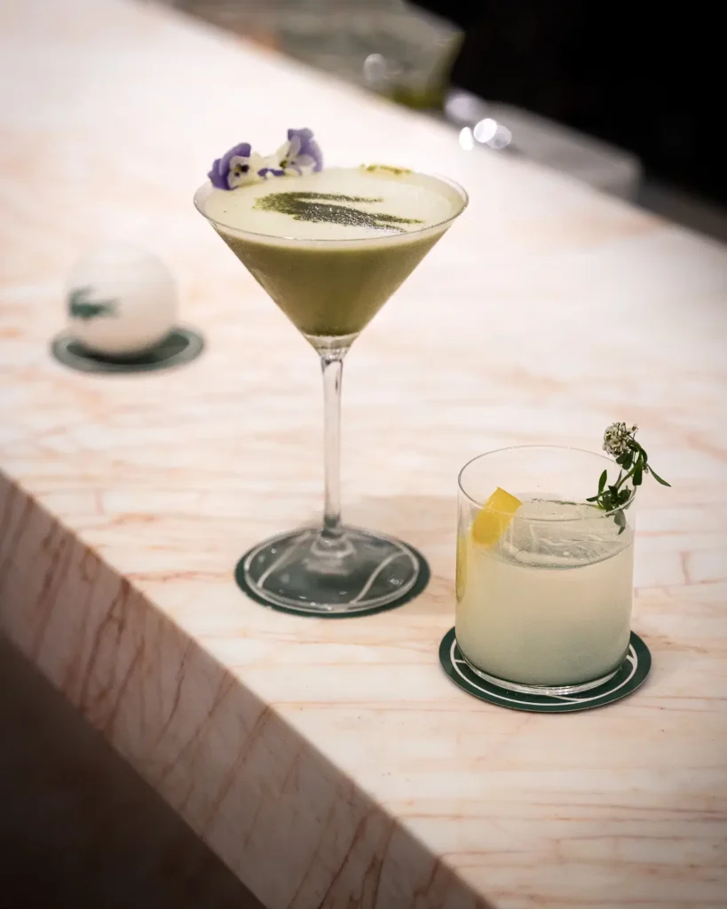Tennis-inspired cocktail presentation featuring a frothy green martini and citrus highball on branded coasters, accompanied by a Lacoste tennis ball and luxury bar style
