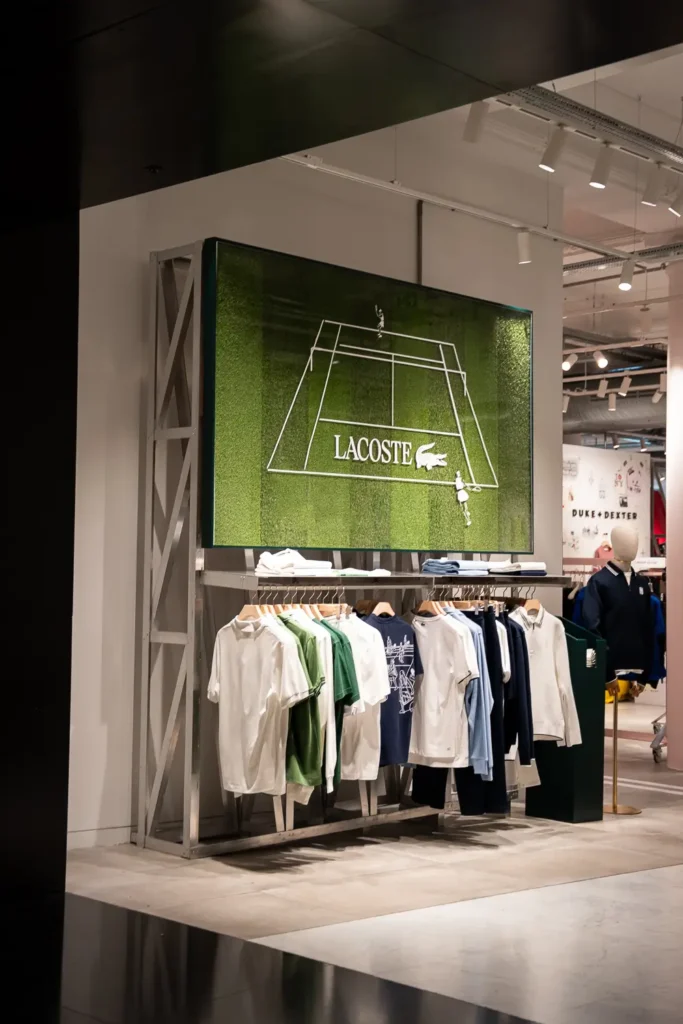 Retail apparel presentation featuring Lacoste tennis-inspired clothing beneath a large grass-court artwork, reinforcing the brand’s deep roots in the sport