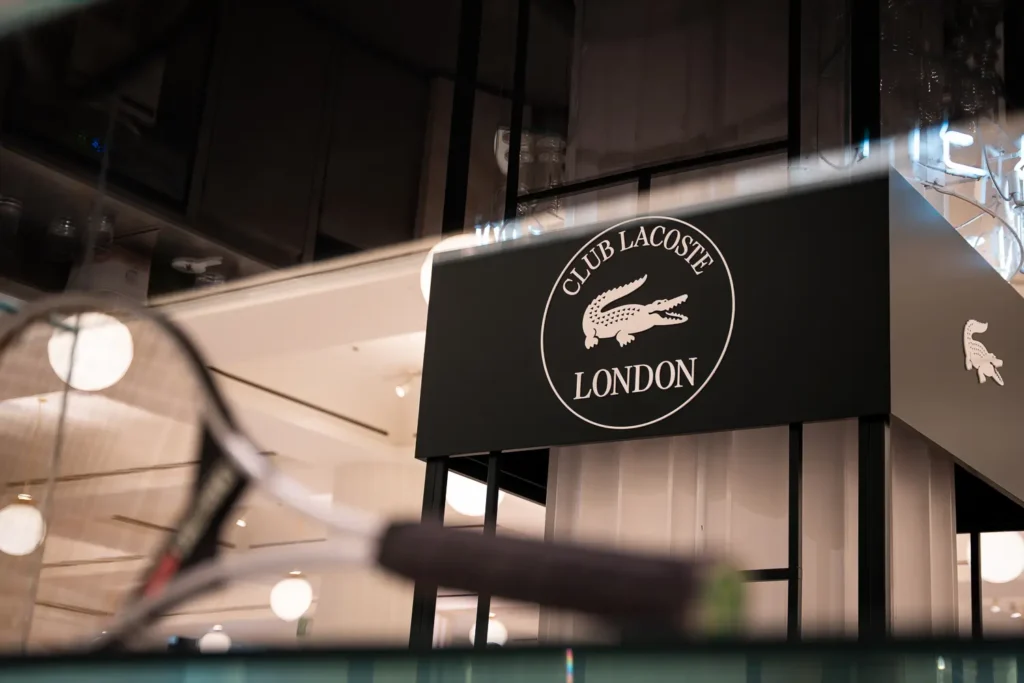 Club Lacoste London illuminated signage framed by blurred tennis rackets and modern retail interiors, emphasizing the brand’s connection to tennis culture and premium lifestyle experience