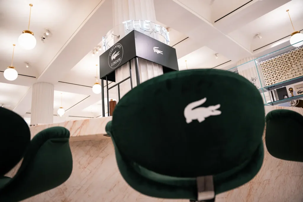 Green velvet bar stools embroidered with the iconic Lacoste crocodile logo surrounding the marble Club Lacoste bar, combining comfort, elegance, and sporting heritage