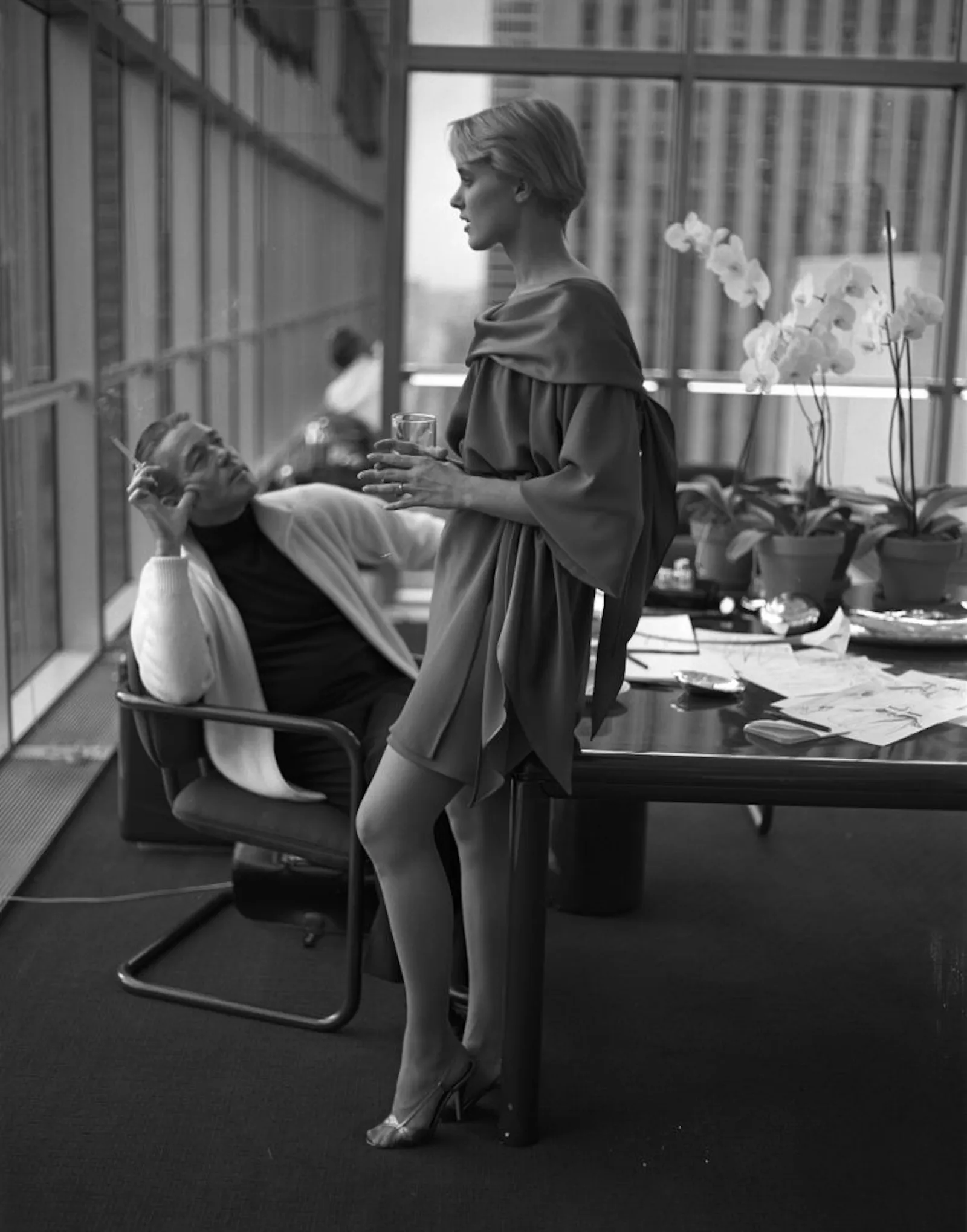 Black-and-white editorial scene in a modern office, featuring a woman in a draped dress standing beside a conference table while a seated man looks up at her. Floor-to-ceiling windows, orchids, and scattered sketches create a refined fashion and creative-studio atmosphere