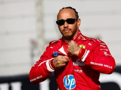 A Formula 1 driver wearing a bright red racing suit adjusts the front of his uniform while standing trackside. Dark sunglasses, braided hair, and visible hand tattoos add to the confident look. The shallow depth of field keeps the focus on the driver, with the racetrack environment softly blurred in the background