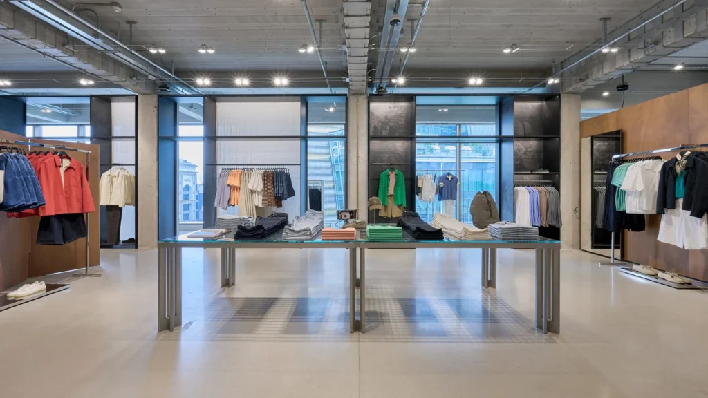 A modern apparel showroom featuring large glass display tables, industrial concrete columns, and expansive windows. Folded garments and color-coordinated clothing collections are arranged with precision, while natural daylight highlights the store’s open, contemporary layout