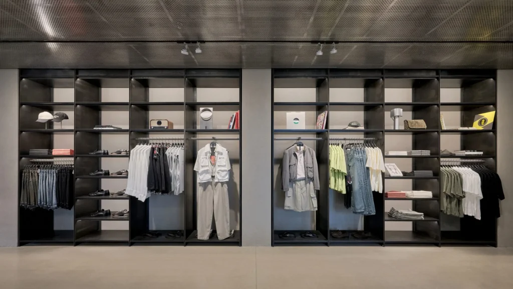 An upscale retail space combining industrial architecture with curated fashion displays. A central glass table presents folded apparel and accessories, while built-in shelving, clothing racks, and decorative objects create a sophisticated boutique environment