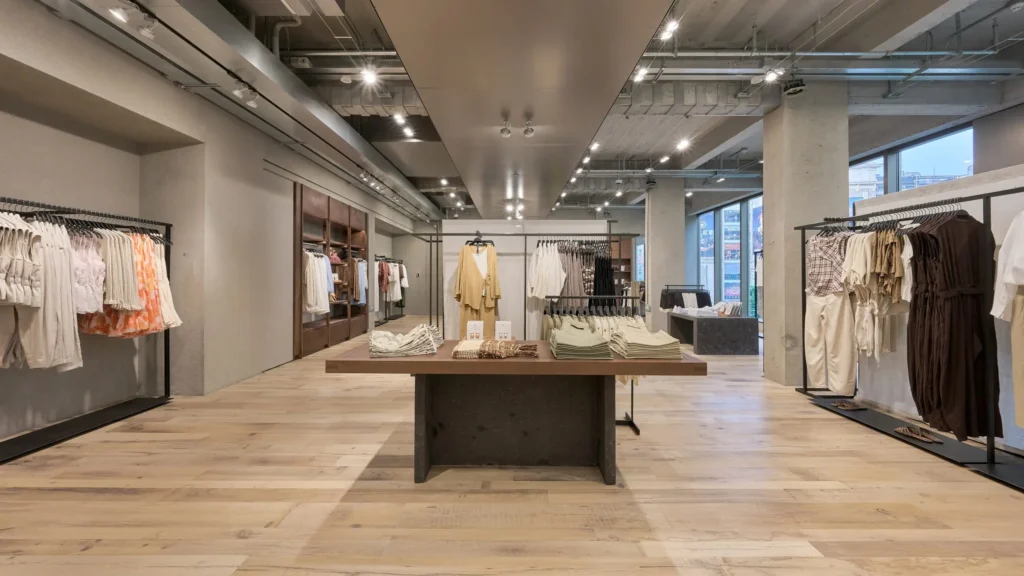 Custom black shelving units line the wall of a modern clothing store, showcasing neatly arranged garments, footwear, books, and accessories. The structured display system balances fashion merchandising with lifestyle elements in a clean, minimalist set
