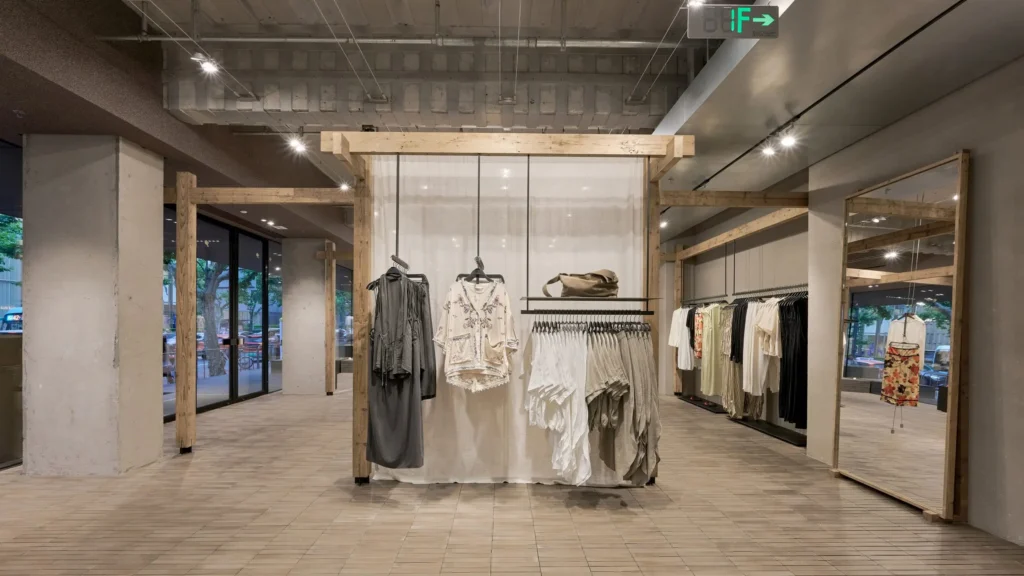 A minimalist retail installation constructed from natural timber frames and soft fabric partitions. Dresses and garments hang within the pavilion-like structure, creating an airy showroom environment that blends fashion presentation with architectural design