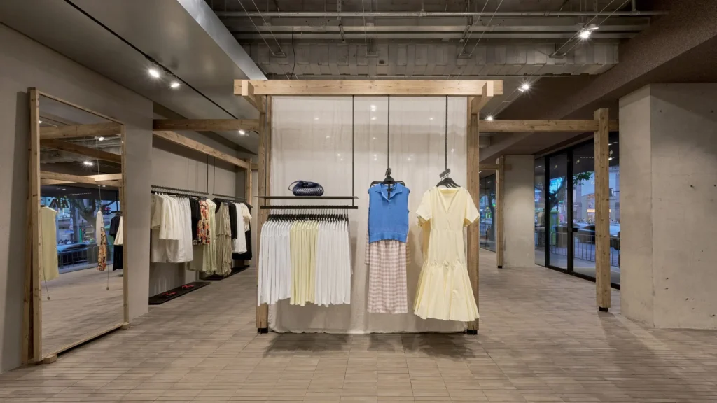 A contemporary fashion boutique featuring timber-framed display structures, suspended garments, and soft neutral textiles. Large mirrors and expansive windows enhance the open layout, while carefully curated apparel creates a relaxed luxury atmosphere