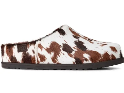 Side profile of a UGG slip-on clog featuring genuine calf hair with a brown, black, and white cow-print pattern. The backless silhouette has a rounded toe, low-profile dark rubber sole, exposed seam at the vamp, and a subtle embossed UGG logo near the heel