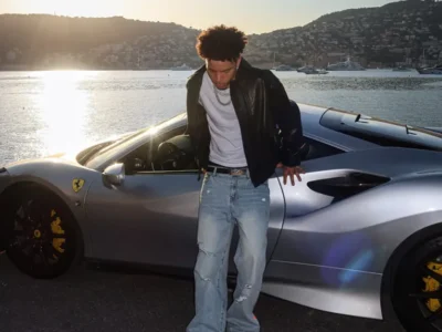 Lil Mosey in a black leather jacket, white T-shirt, and light-wash jeans stands beside a silver Ferrari at a waterfront marina during sunset. A "Parental Advisory" label appears in the bottom-right corner