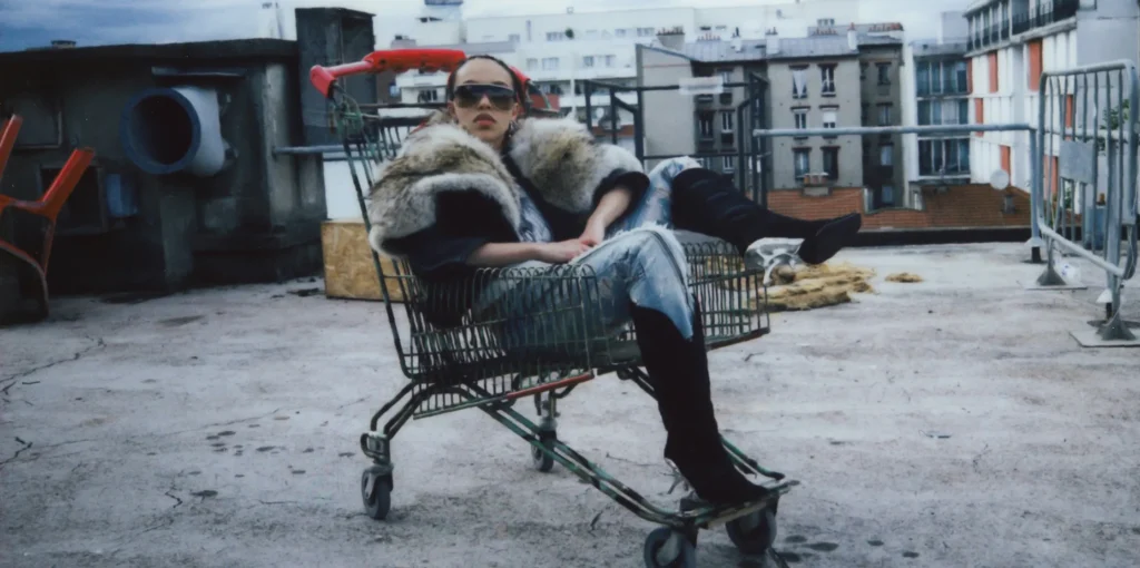 Fashion editorial photograph of a model reclining inside a shopping cart on an urban rooftop. The model wears oversized sunglasses, a dramatic fur-trimmed coat, metallic silver trousers, and black knee-high boots. Apartment buildings and industrial rooftop elements create a moody, cinematic backdrop with a grainy, film-inspired