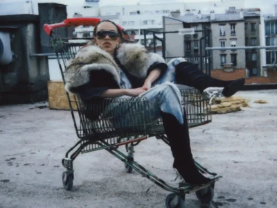Fashion editorial photograph of a model reclining inside a shopping cart on an urban rooftop. The model wears oversized sunglasses, a dramatic fur-trimmed coat, metallic silver trousers, and black knee-high boots. Apartment buildings and industrial rooftop elements create a moody, cinematic backdrop with a grainy, film-inspired
