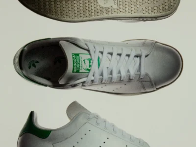 Vintage-inspired adidas Stan Smith 80s sneaker shown from outsole, top-down, and lateral views, featuring premium white leather construction, green heel tab accents, perforated Three Stripes branding, and an aged off-white sole unit
