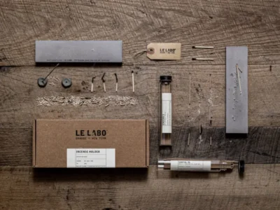 Le Labo home incense collection displayed in a flat-lay arrangement on a rustic wooden surface, featuring incense sticks, a minimalist incense holder, branded packaging, glass fragrance tubes, and handcrafted materials arranged with precise, workshop-inspired detail